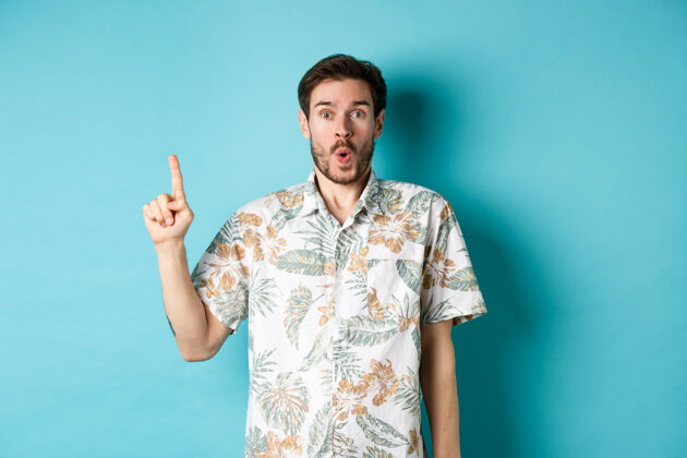 朋友惊讶的白人游客穿着夏威夷衬衫 指着手指说哇哦 查看促销活动 站在蓝色背景上自信成年人酷
