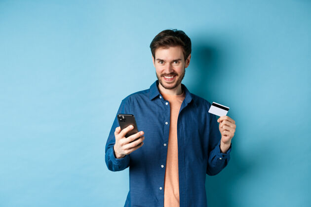 在线电子商务概念英俊的年轻人在网上购物 拿着智能手机和塑料信用卡 对着镜头微笑 站在蓝色的背景上小玩意胡须站立