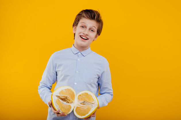热带笑容满面的小男孩手里拿着柚子 穿着蓝色衬衫 隔离在黄色的墙上积极维生素小