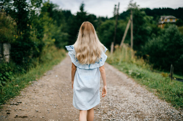 道路年轻迷人优雅的金发碧眼女孩 身着蓝色连衣裙 在乡间的马路上摆姿势乡村背部年轻