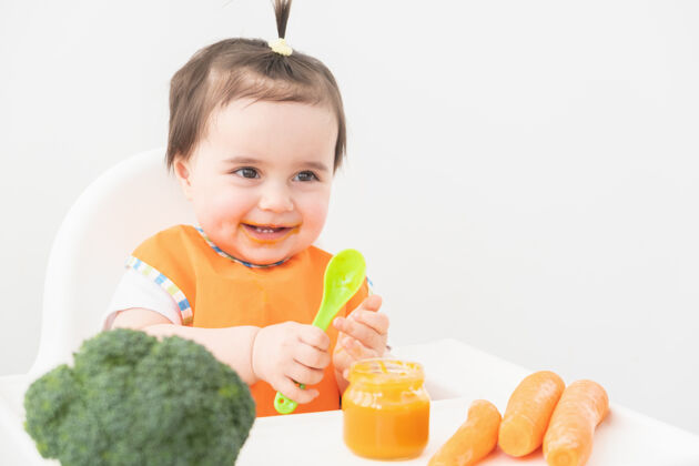 花椰菜穿着橙色围嘴的女婴坐在儿童椅上吃白色背景的蔬菜泥保健母亲胡萝卜