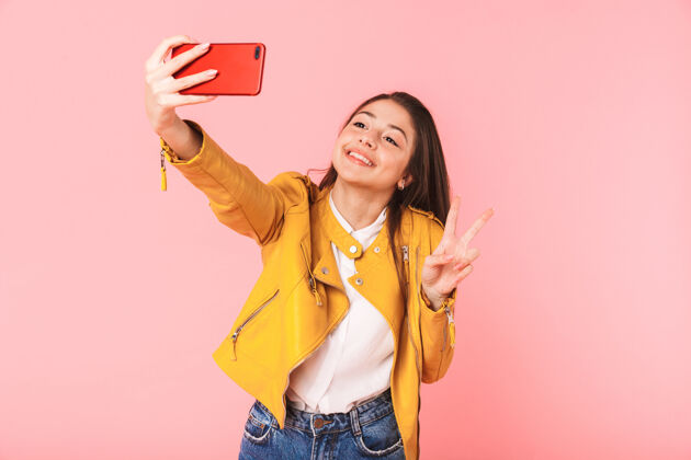 Take美丽的年轻女孩在粉红色的墙上摆出孤立的姿势 用手机自拍手机童年肖像