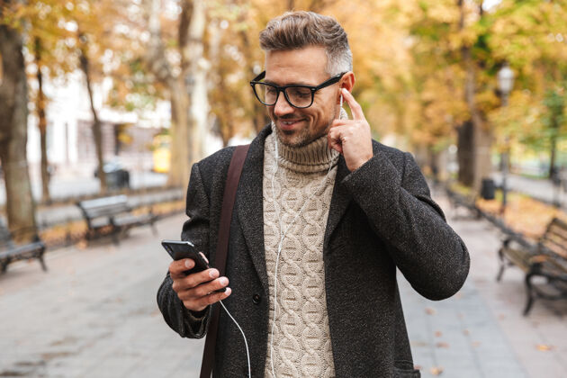 手机帅哥穿着外套走在户外 用耳机和手机听音乐优雅户外音乐