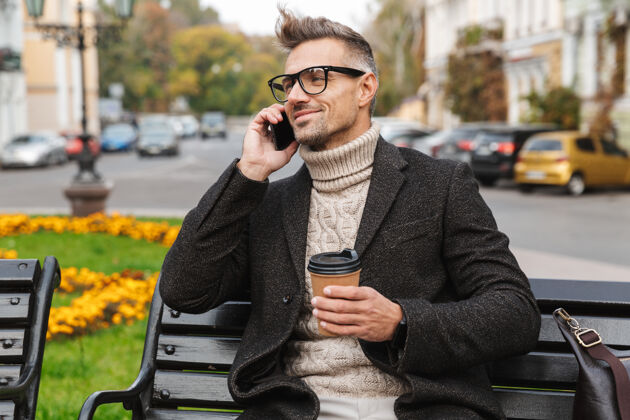 男士穿着外套的帅哥坐在户外的长凳上 讲着手机 手里拿着外卖咖啡杯忙碌帅气通话