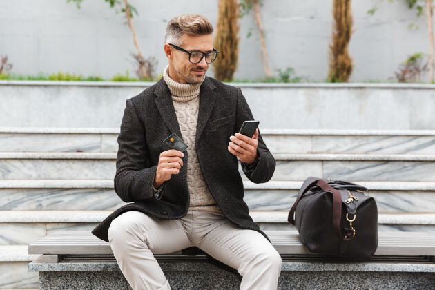 男孩帅哥穿着夹克坐在户外用手机和信用卡上网购物休闲买家网络