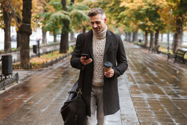 胡须图为30多岁的快乐男人穿着暖和的衣服 在户外穿过秋天的公园 并使用手机手机寒冷公园
