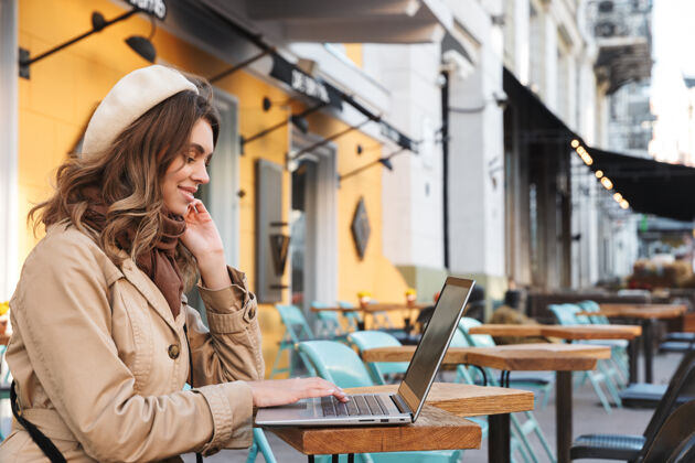 秋天穿着外套的漂亮年轻女子坐在咖啡馆外面用笔记本电脑打字短信肖像