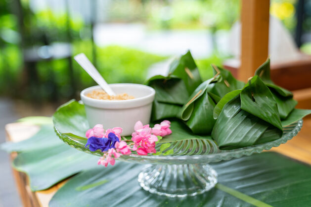 碗泰国传统的甜点在香蕉叶里面包上玻璃盘 吃的时候旁边放糖它安排在室外花园的木桌上户外天然美食