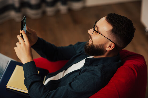 手机带着微笑的年轻胡须戴着眼镜的男子正在使用智能手机他看有趣的视频沙发男性现代人