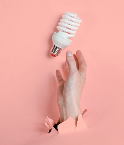手臂女性手拿着螺旋灯泡穿过撕破的粉红色纸简约创意时尚理念灯能源环境