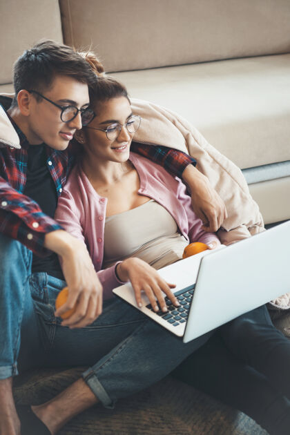 家庭一对年轻可爱的夫妇坐在沙发旁边的地板上 一边看笔记本电脑上的东西 一边吃被子盖着的橘子真实手臂关系