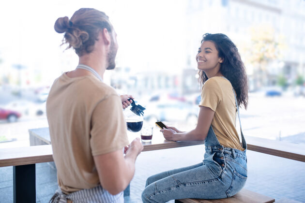 千禧一代时间到了咖啡快乐一个穿着牛仔工作服的女人坐在咖啡馆靠近大窗户的地方 一个穿着围裙的男人拿着一壶咖啡黑头发男人金发