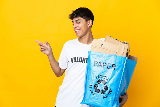 微笑一名男子拿着一个装满纸的回收袋 在孤立的黄色背景上进行回收 手指指向侧面 展示一个产品指向保护垃圾