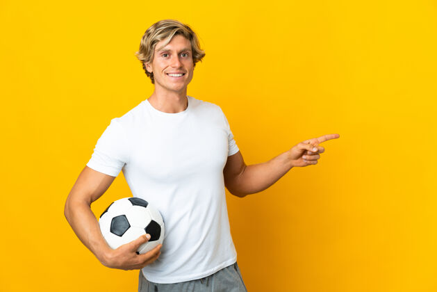自信在孤立的黄色背景上的英国足球运动员用手指指着侧面足球介绍金发