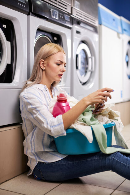 家务在洗衣房里 一名女性坐在地板上 靠在洗衣机旁 手里拿着一部智能手机豪斯金发白种女人在等着呢无聊卫生女人
