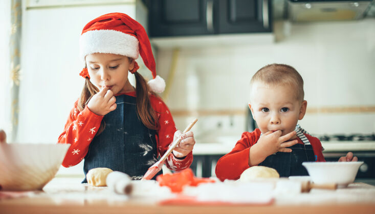 衣服高加索兄弟姐妹穿着圣诞老人的衣服在厨房为圣诞节准备食物房子帽子蛋糕