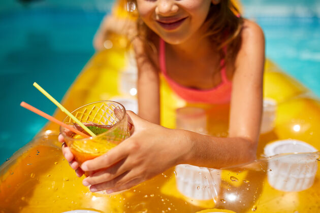 小小女孩在游泳池里放松 享受日光浴 在家庭度假时在水里的黄色充气床垫上喝果汁床垫鸡尾酒休闲