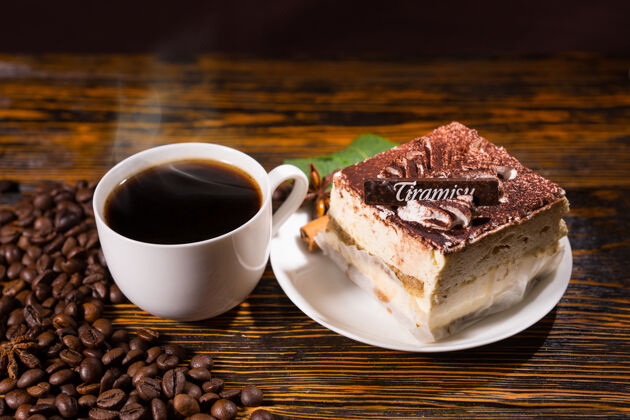 标签美味的方形蛋糕片放在盘子里 旁边是一整杯咖啡 周围是深色的咖啡豆 背景是染色的木头提拉米苏美味写作