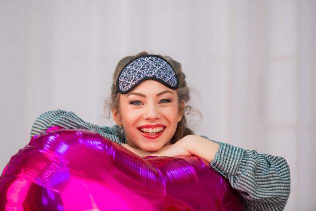 可爱一个戴着睡眠面罩的健康女孩微笑着拥抱着一个心形的粉色气球床年轻蓝眼睛