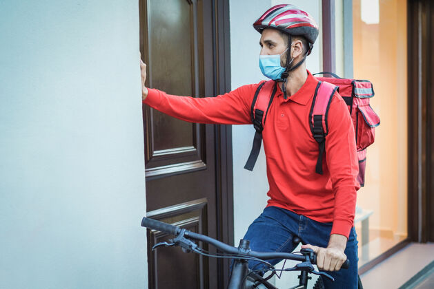 面具年轻的快递员背着热背包 骑着自行车按着门铃-注意脸防护年轻健康