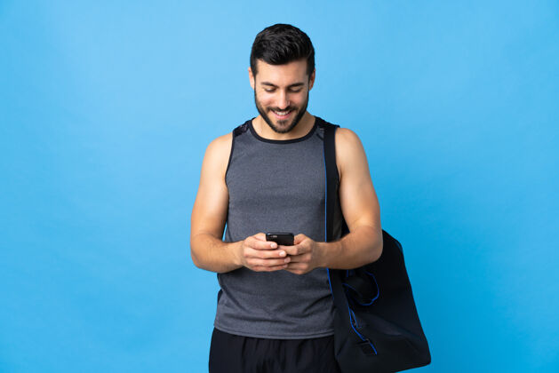 说话年轻的运动男子与运动包孤立的蓝色发送信息与手机科技胡须移动