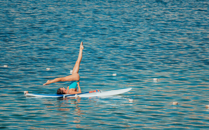 比基尼穿着蓝色泳衣的年轻女子用桨在冲浪板上做瑜伽湖泊早晨成人