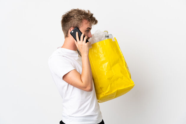 青年年轻的金发男子拿着一个装满废纸的回收袋 在白色的地板上和别人用手机交谈男性再利用生态系统
