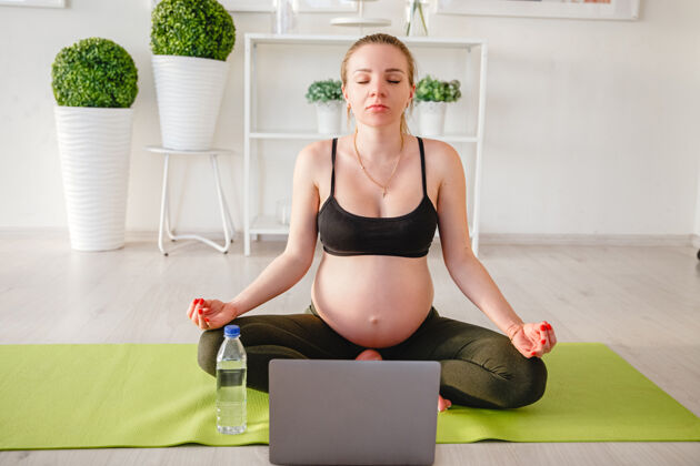 电脑年轻的怀孕的金发女人在家里的垫子上做瑜伽笔记本电脑高高质量的照片坐着室内怀孕