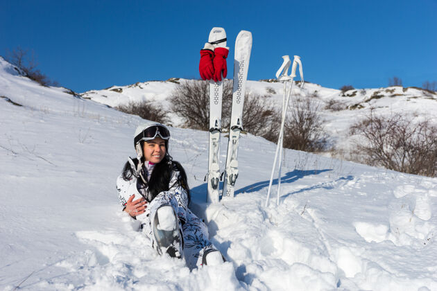 峰会在晴朗的天空和温暖的阳光下 年轻女子戴着头盔和护目镜 坐在雪地里 黑色长发 滑雪板和杆子就在附近极点冬季天空