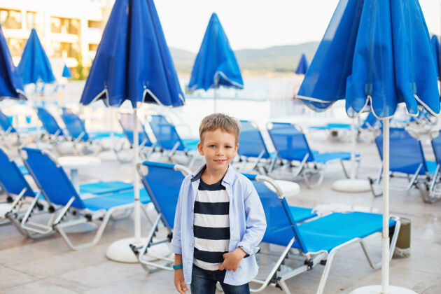 椅子在一个避暑胜地 一个小男孩站在蓝色雨伞和日光浴者中间放松脸水