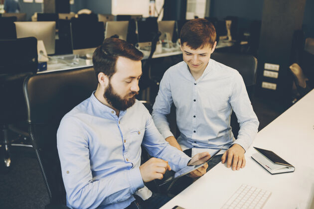 同事两个穿着衬衫的男人坐在时髦的办公室里 一边用现代的平板电脑一起男同事在办公桌上浏览平板电脑办公室在线专业