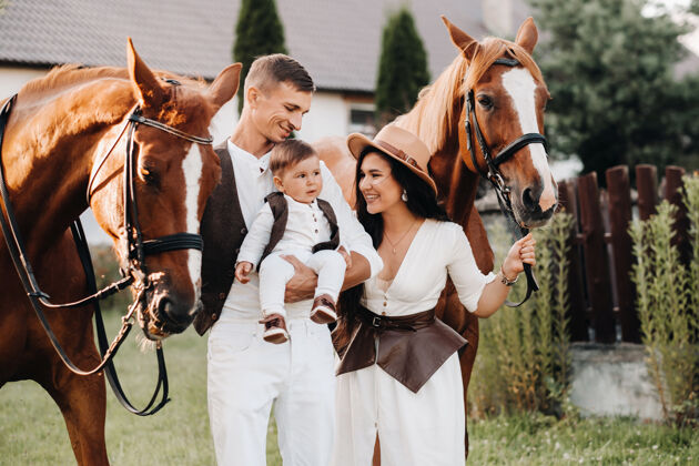 幼儿一个穿着白色衣服的家庭和他们的儿子站在大自然中两匹美丽的马旁边一对时髦的夫妇带着一个孩子与马合影快乐动物日落