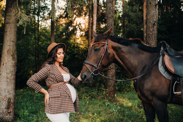 动物一个大肚子的怀孕女孩戴着帽子在森林里的马旁边自然时尚穿白色衣服和棕色夹克的女孩夜晚户外快乐
