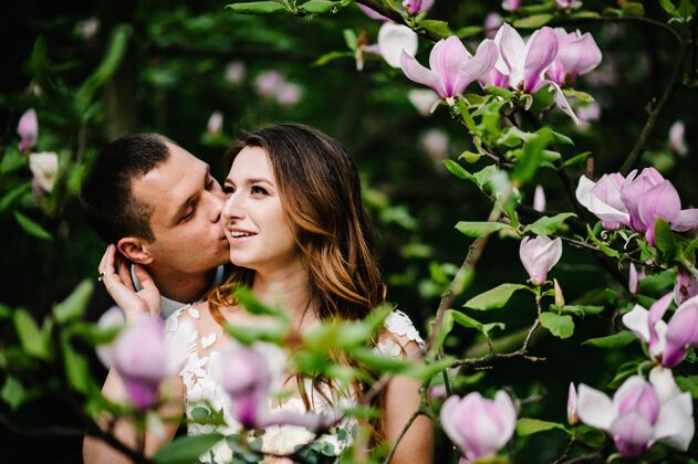 婚礼一对新人在公园里拥抱亲吻 背景是粉色和紫色的玉兰花格林婚礼仪式地点自然树花园