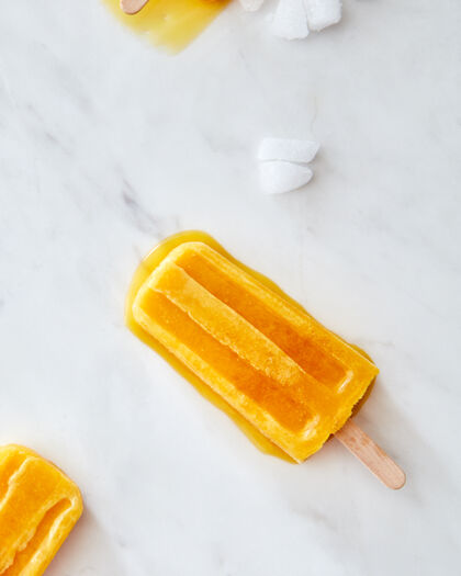 冰霜橙色水果冰激凌在一根棍子上 在灰色大理石背景上放上几片冰 留出空间文本夏季概念甜点平底躺下水果自制棒棒糖汽水