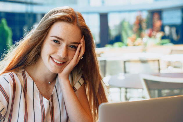 肖像一位红发雀斑的可爱年轻女子的画像 她微笑着用手触摸脸 在咖啡馆的户外笔记本电脑上工作会议自由空间餐厅