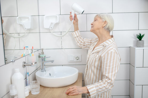 浴室做头发灰色-一位头发花白的老妇人在浴室里吹干头发养老金每日吹风机