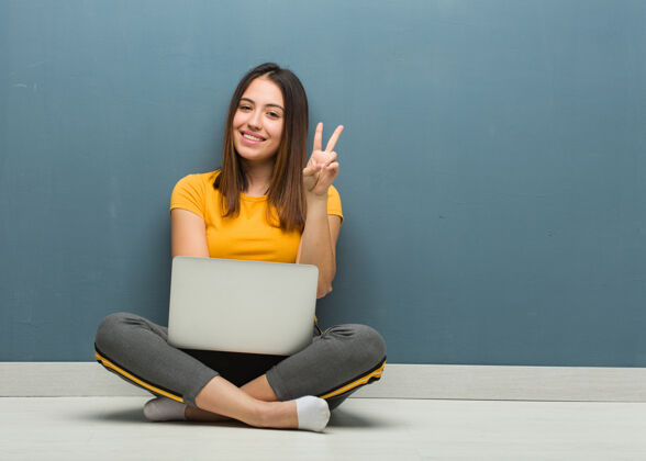 女性坐在地板上拿着笔记本电脑的年轻女子 上面显示着第二个计算机指向胜利