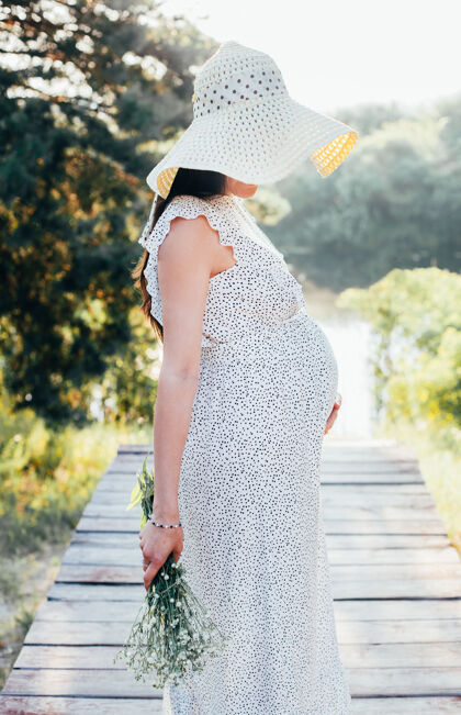 人一个怀孕的女孩 戴着一顶夏天的帽子 穿着圆点太阳裙 在树的背景上放着一束雏菊洋甘菊太阳裙树
