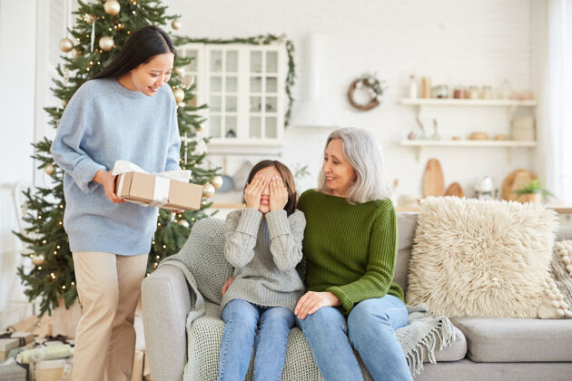 姐姐微笑的女人给她的小妹妹圣诞礼物 而他们坐在沙发上庆祝圣诞节礼物家里圣诞树