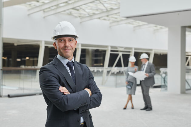 工作微笑的成熟商人戴着安全帽 双手交叉站在工地上的腰间画像 承包商场地建筑