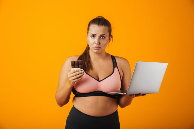 白种人一个穿着运动服的失望的超重年轻女子的肖像 孤立地站在黄色的墙上 用笔记本电脑 拿着巧克力棒活动幸福运动
