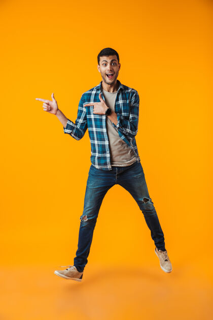 男一个穿着格子衬衫的快乐的年轻人的全长肖像被隔离在橙色的墙上 走着 指着别处健身年轻男