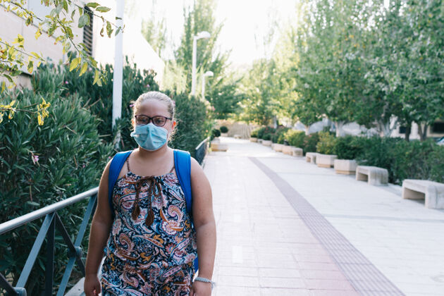 冠状病毒一个戴着眼镜 蓝色背包和面罩的金发女孩在上学的路上听力学生流行