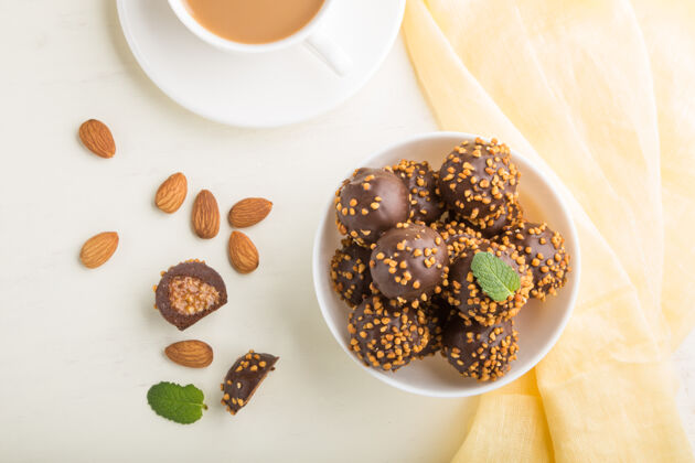盘子巧克力焦糖球糖果杏仁和一杯咖啡在一个白色的木制表面和黄色的纺织品杏仁杯子叶子