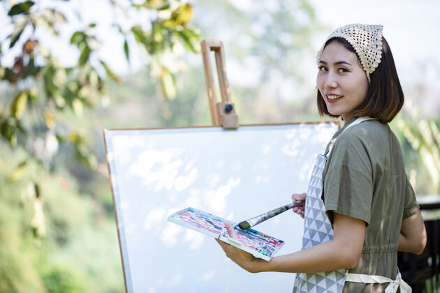 画笔夏日田野里的亚洲美女 用画笔和彩色颜料在花园里的纸艺板上画画休闲美丽作坊