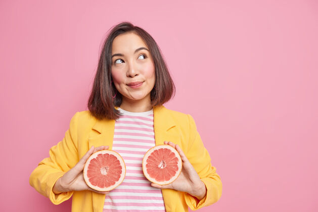 思想迷人的亞洲女人有著深思熟慮的夢幻表情抱著葡萄柚一半吃多汁的柑橘類水果燃燒熱量有著健康的營養姿勢靠著粉色的墻面和復制空間區思維切片沉思