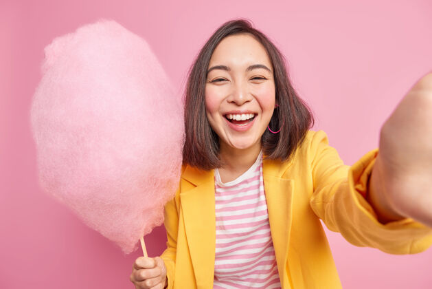 乐观快乐真诚的亚洲女人表达真实的情感拿着美味的糖果牙线积极地自拍微笑夏天散步时心情好穿着时尚的衣服隔离在粉色的墙上黑发糖棉花
