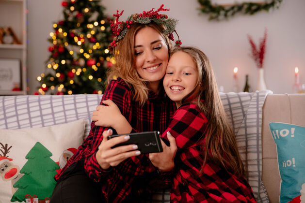 时间高兴的母亲和冬青花环和女儿看电话坐在沙发上 享受圣诞节的时间在家里享受房子沙发