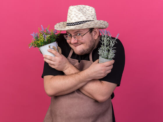 人贪婪的年轻男园丁戴着园艺帽 拿着和粉色墙壁隔离的花盆里的花在一起持有交叉帽子